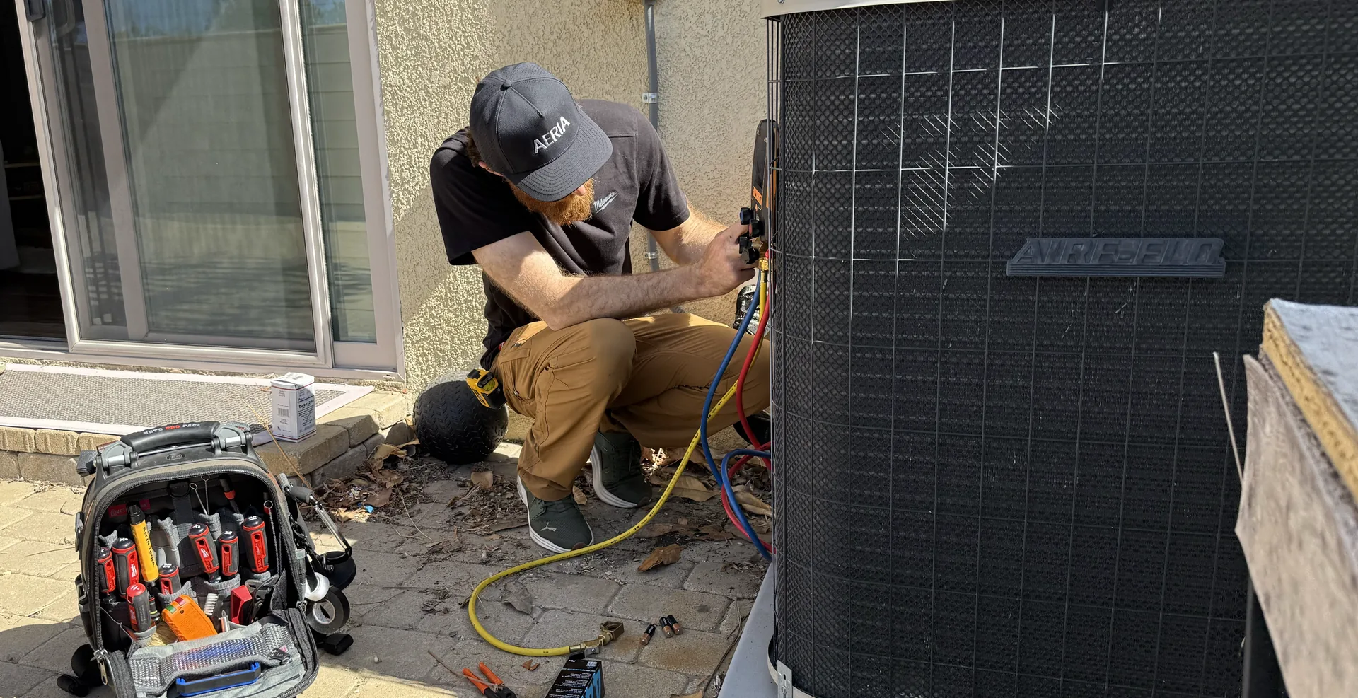 Technician in an AERIA cap connecting gauge hoses to a residential outdoor AC unit, with an open tool backpack on the patio