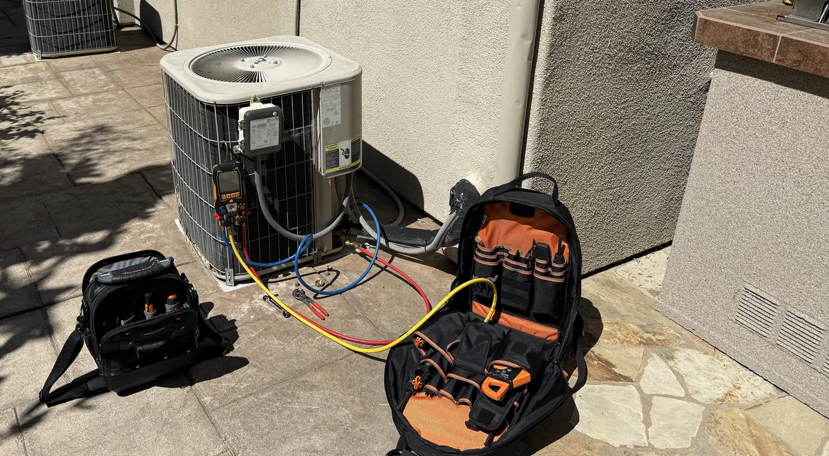 Digital refrigerant manifold and hoses connected to a residential outdoor AC unit, with two open tool bags on the patio