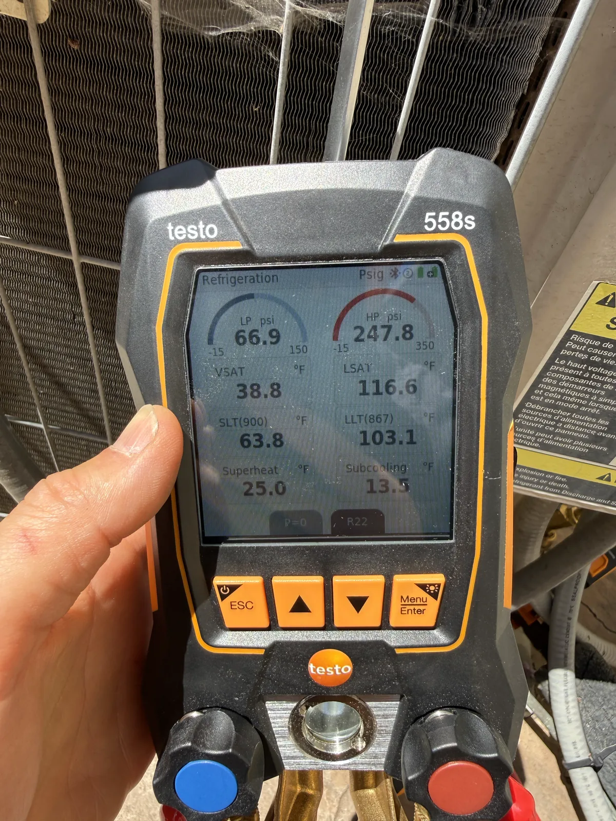 Hand holding a digital refrigerant manifold that displays live system pressures and temperatures beside an outdoor AC coil
