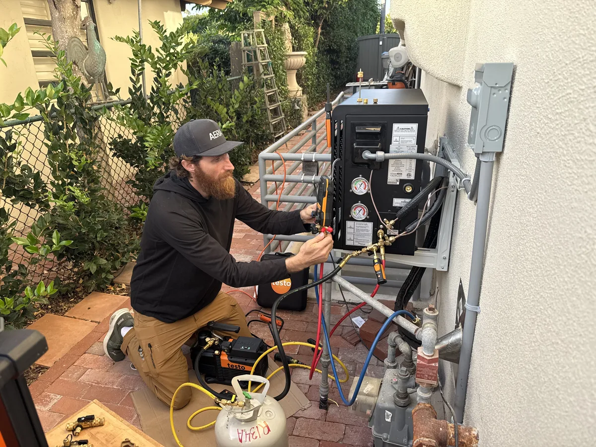 AERIA technician kneeling at a wall-mounted condensing unit with a manifold, vacuum pump and refrigerant cylinder connected
