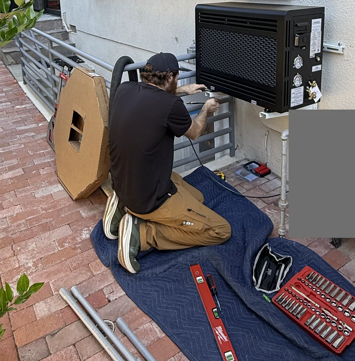 Technician kneeling on a padded blanket laid over brick pavers, with a socket set and level beside wall-mounted equipment