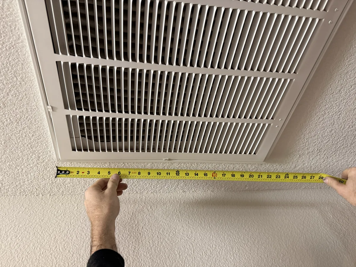 Two hands holding a tape measure alongside a ceiling return-air grille