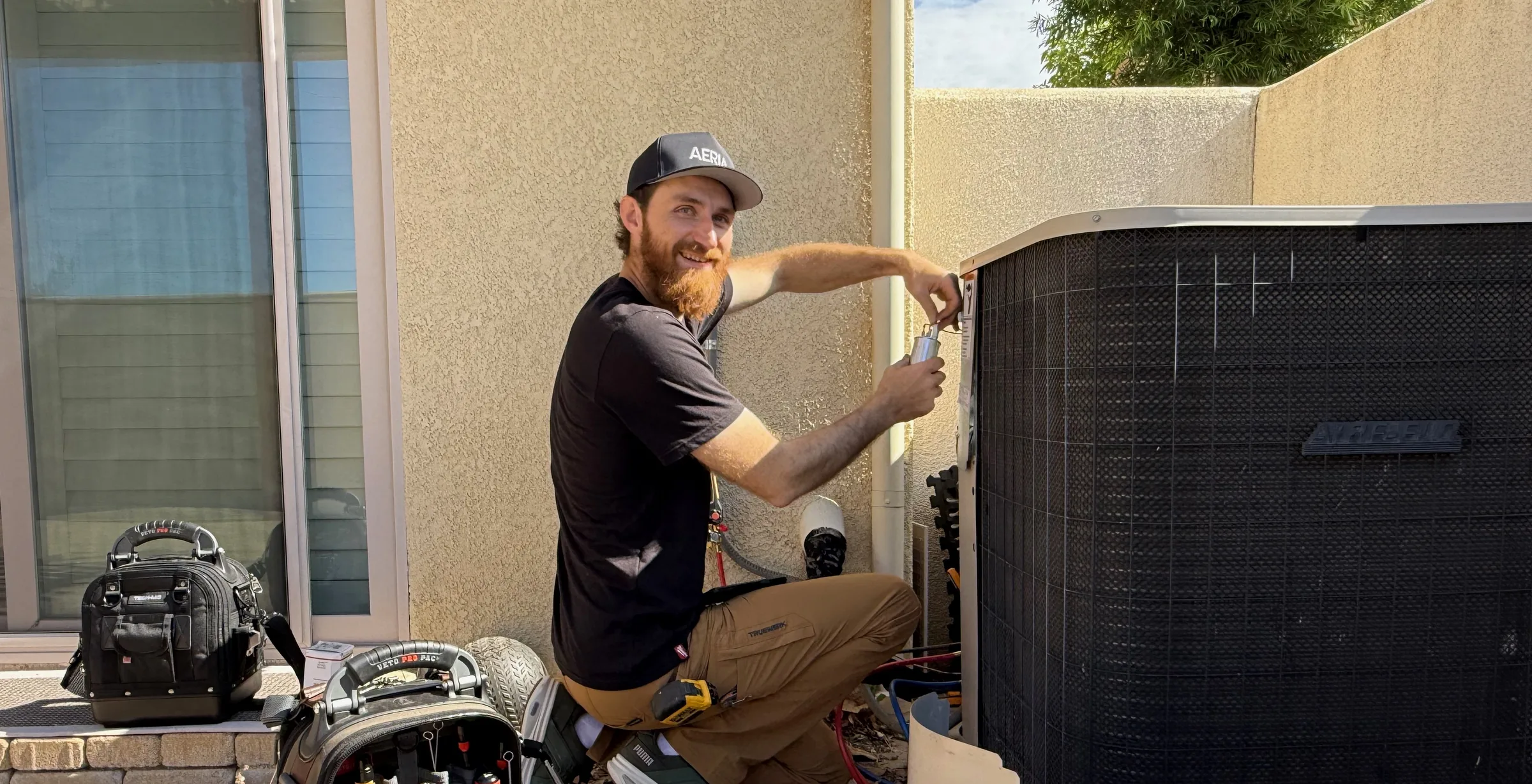 AERIA technician kneeling at the open service panel of a residential outdoor AC unit, with tools laid out on the patio
